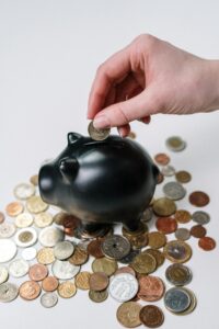 Hand inserting coin into a black piggy bank surrounded by various coins, symbolizing savings.